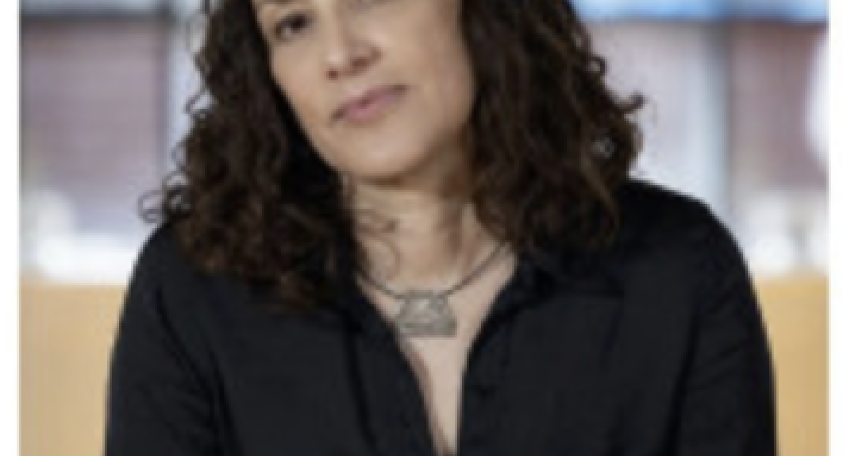 Silvana Cardell is a Latina woman with light skin and dark, shoulder-length curly hair. She wears a black button-down shirt and a simple necklace. Seated and facing the camera, she has a calm expression in soft, even light.