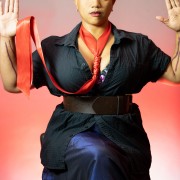 Photo of a light skinned black femme in a black button up shirt and dark blue long skirt holding their hands up with a soft red noose as a necklace