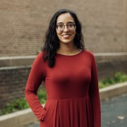A Latinx cis woman with wavy hair and smiling, wearing purple glasses and a red dress