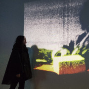 A person stands in a dimly lit room, facing a large, grainy video projection on the wall. The projection shows an abstract, colorful image in tones of yellow, green, red, and gray. The person’s silhouette is partially illuminated by the light from the screen, creating a contemplative atmosphere.