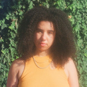 a lightskinned agender person with their big curly hair out wearing a yellow shirt and two chain necklaces in front of a wall covered in ivy