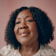 Brown skin women with curly hair and a slight smile, wearing a gold necklace and cream and brown top. 
