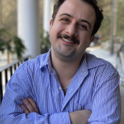 Ozma, a white man in his 30s with curly hair and a mustache, crosses his arms and smiles into the camera. He wears a striped blue button-up shirt.