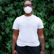 portrait of Black agender person in an N95 mask. they're wearing a white short sleeved t-shirt, tortoiseshell glasses, black pants, and are standing in front of a wall of english ivy