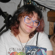 Photo of CJ Weintraub smiling, a white femme with brown and red hair, wearing blue glasses and a white shirt