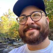 Mid-30s white man with a beard wearing a baseball cap and round glasses, standing in front of a waterfall.