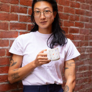 East Asian person with black wire glasses and shoulder-length black hair looks at camera. Standing against a brick wall, they wear a white shirt, and hold up a small mug.