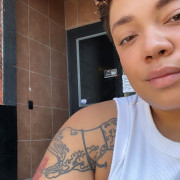 Image of artist Nia Benjamin. Their body is cropped slightly out of frame, they are wearing a white tank top and tattoos on their left arm are visible in the image. 