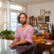 Imran Siddiquee sits at a kitchen counter in a lavender dress smiling at the camera, behind them is a living room in their apartment with various art pieces on the wall visible.