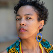 Raina León looks at the side at the camera, wearing blue and yellow beaded earrings, red and yellow beaded necklace, and turquoise and silver necklace, and a printed best