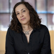 Silvana Cardell is a Latina woman with light skin and dark, shoulder-length curly hair. She wears a black button-down shirt and a simple necklace. Seated and facing the camera, she has a calm expression in soft, even light.