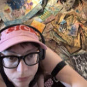 46 year old female identifying person wearing a pink hat with "sick" embroidered on it in red. In the background, colorful sculpted paintings are strewn over the ground.