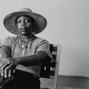 A black and white photo of angel, a brown skinned person wearing a wide brimmed hat and grey top with a long skirt. their legs are crossed on their hands placed lightly on their knee. 