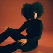 A person with a large natural afro sits on the floor against a warm orange background, wearing a fitted black long-sleeve turtleneck and pants, lit dramatically with shadows across their face and body.