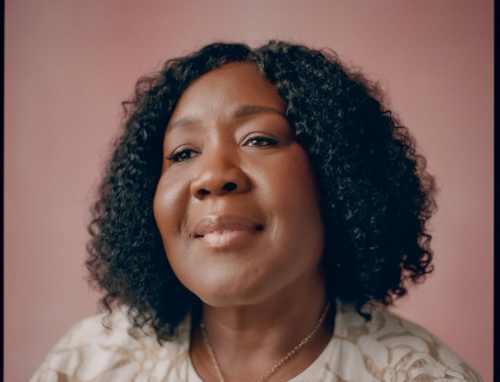 Brown skin women with curly hair and a slight smile, wearing a gold necklace and cream and brown top. 