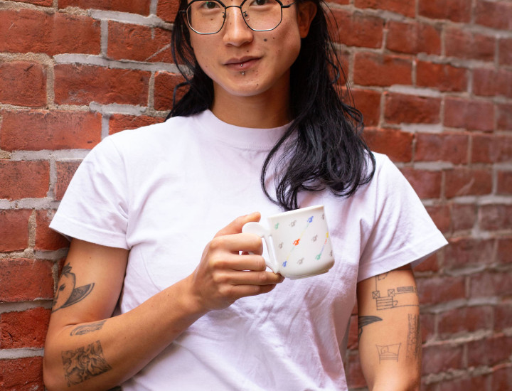 East Asian person with black wire glasses and shoulder-length black hair looks at camera. Standing against a brick wall, they wear a white shirt, and hold up a small mug.