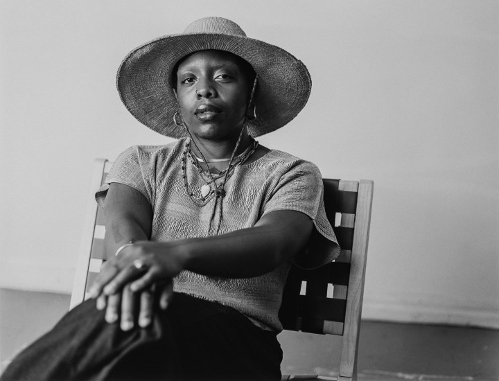 A black and white photo of angel, a brown skinned person wearing a wide brimmed hat and grey top with a long skirt. their legs are crossed on their hands placed lightly on their knee. 